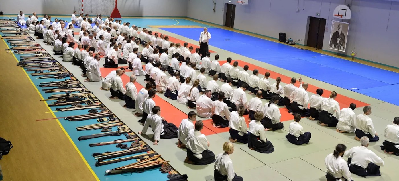 Vue d'ensemble d'un stage : pratiquants assis en seiza, armes en bois alignées au bord du tatami