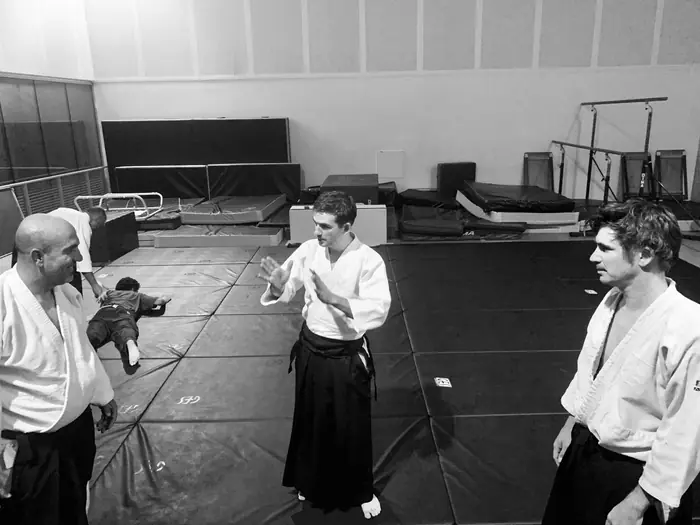 Vue large du dojo pendant un cours, plusieurs pratiquants debout
